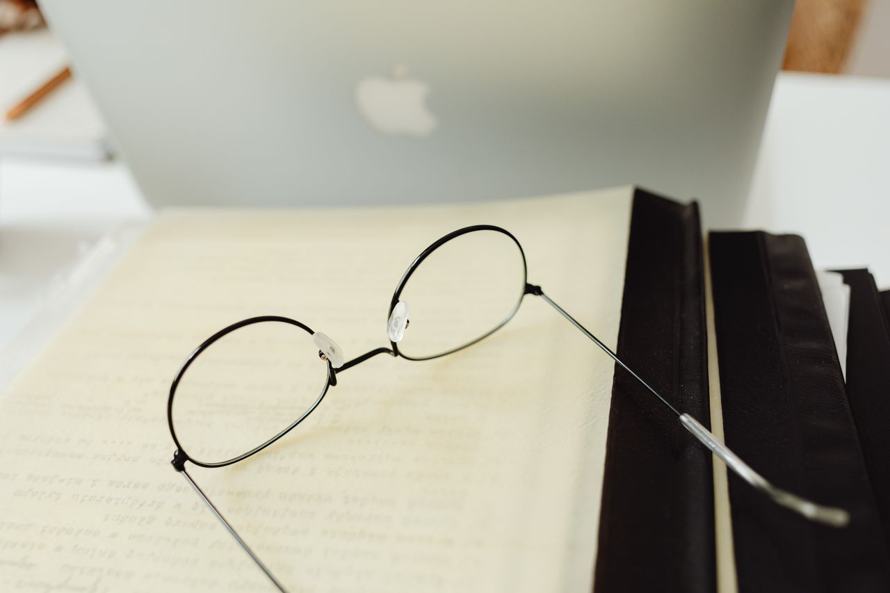 our-services-03 Round eyeglasses resting on an open book beside a laptop in a quiet study atmosphere.